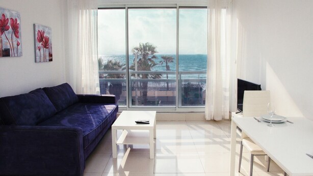 A modern living room with a large window, a blue sofa, a white coffee table, and a dining area with a table and chairs. - GK Apartments - Ben Gurion 105 (Bat Yam)