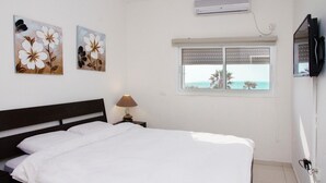 A bedroom with a bed, a nightstand with a lamp, two floral paintings, a window with a view of the sea, and a mounted TV. - GK Apartments - Ben Gurion 105 (Bat Yam)