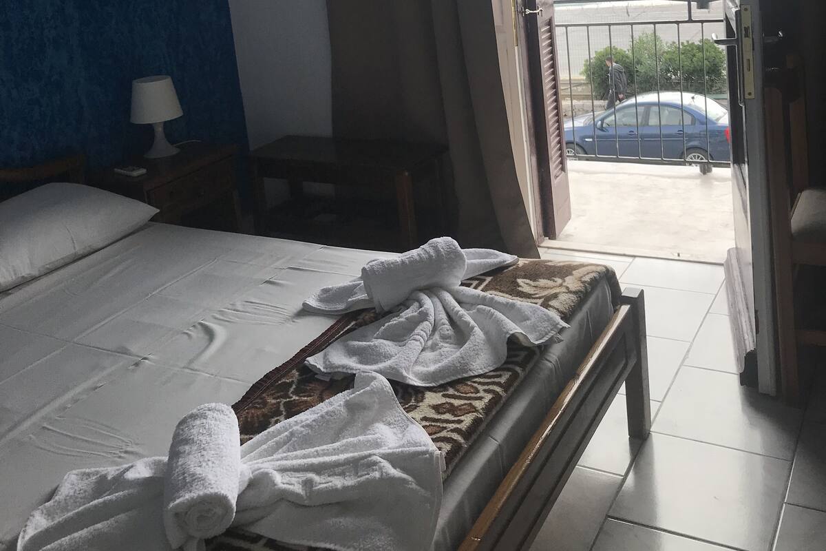 A hotel room with a bed, a bedside table with a lamp, a window with curtains, and a view of a body of water and a boat.