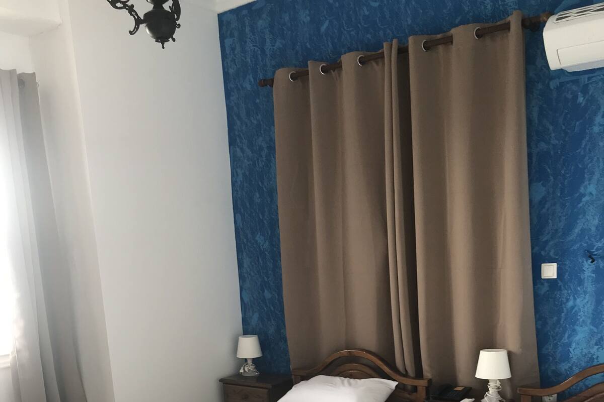 A hotel room with two beds, each with a blue and white bedspread, and a brown curtain. There are bedside tables with lamps and a wall-mounted air conditioner.