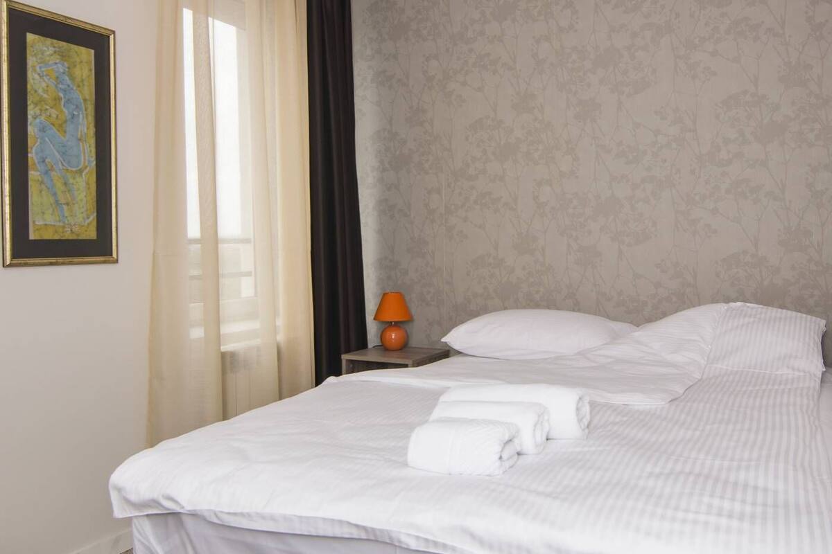 Un lit bien fait, avec du linge de lit blanc, une base de lit grise et une petite table de chevet avec une lampe.