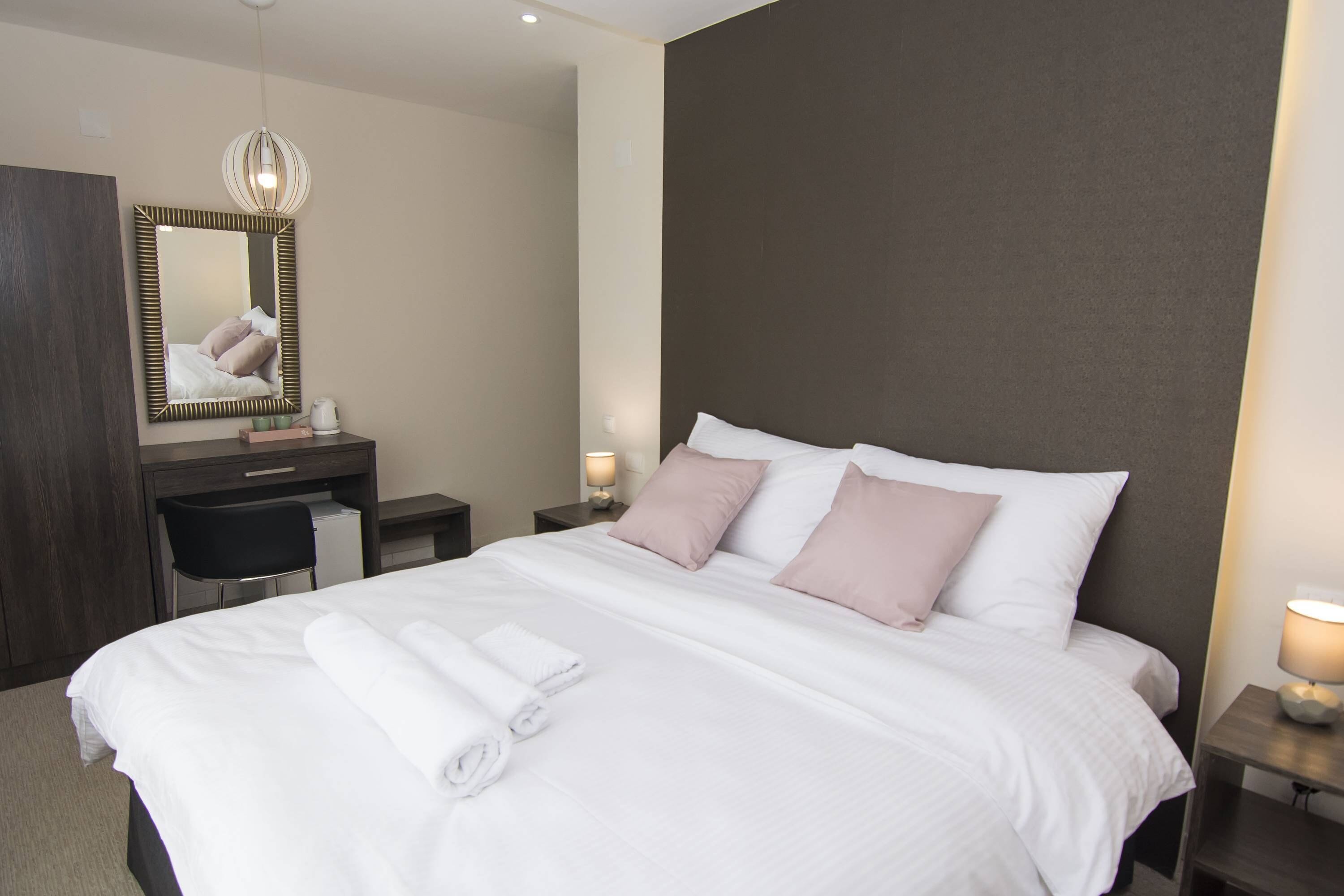 A neatly made bed with pillows, a bedside table with a lamp, a desk with a chair, and a mirror on the wall.