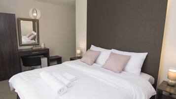 A neatly made bed with pillows, a bedside table with a lamp, a desk with a chair, and a mirror on the wall.