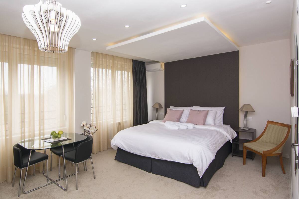 Une chambre d’hôtel moderne avec un grand lit, une table en verre avec des chaises, un bureau et un lustre décoratif.
