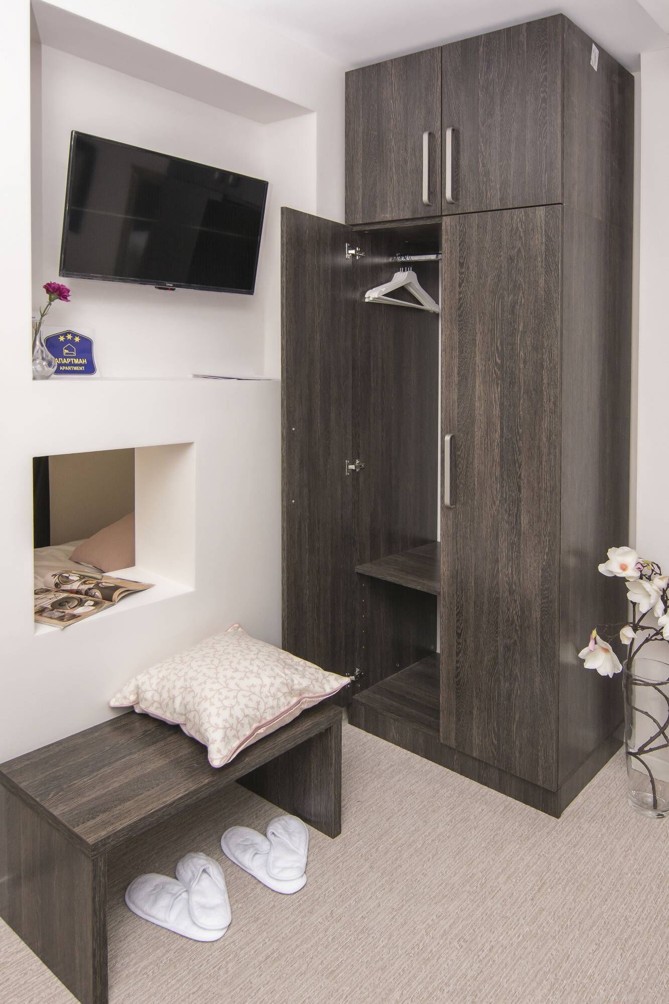 Ein Schlafzimmer mit einem dunklen Holzschrank, einem flachbildfernseher an der Wand, einem kleinen Nachttisch mit einem Kopfkissen und einem Paar Hausschuhen auf dem Boden.