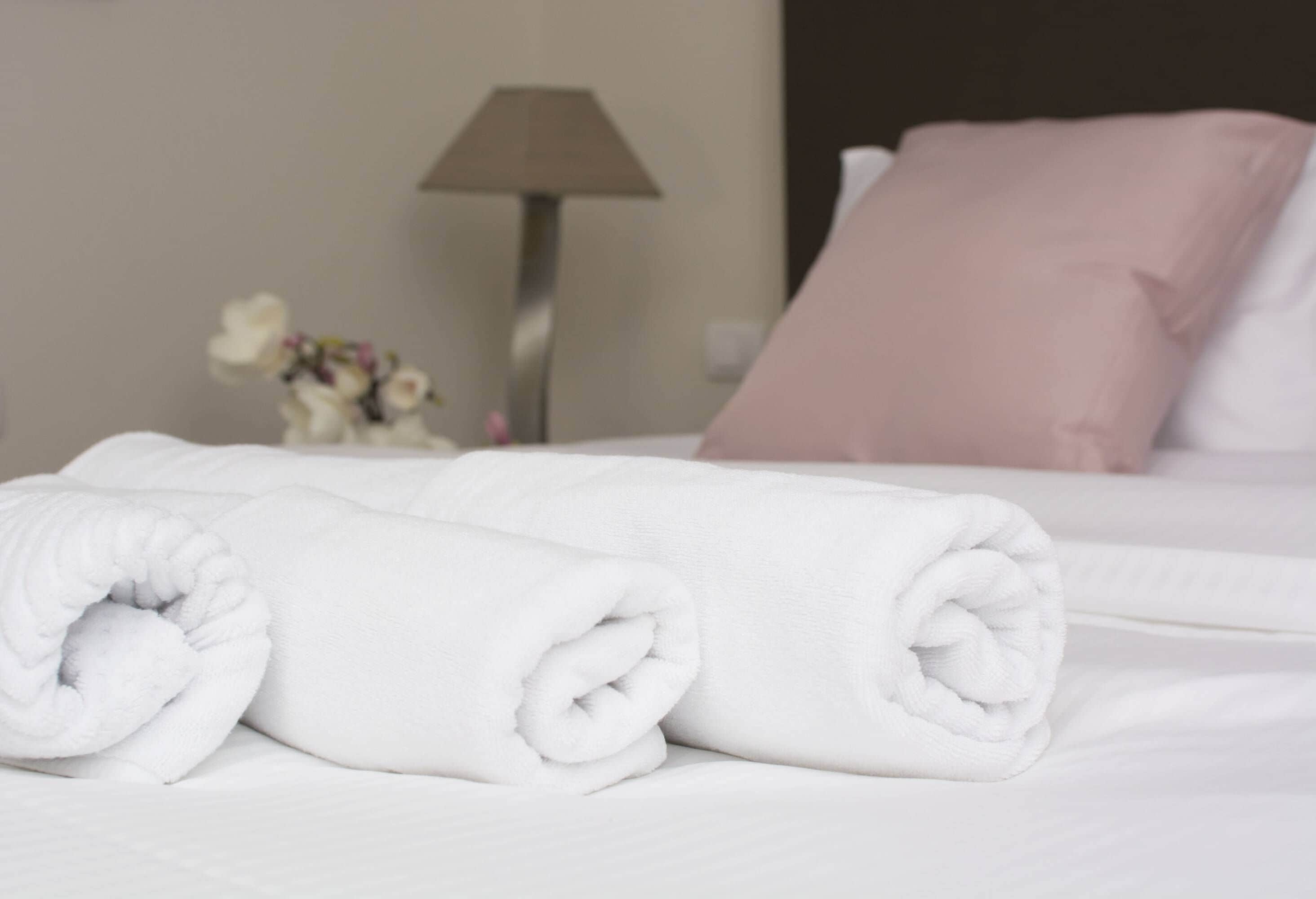 Una cama bien hecha con almohadas blancas y rosas, una toalla blanca enrollada y una lámpara al fondo.