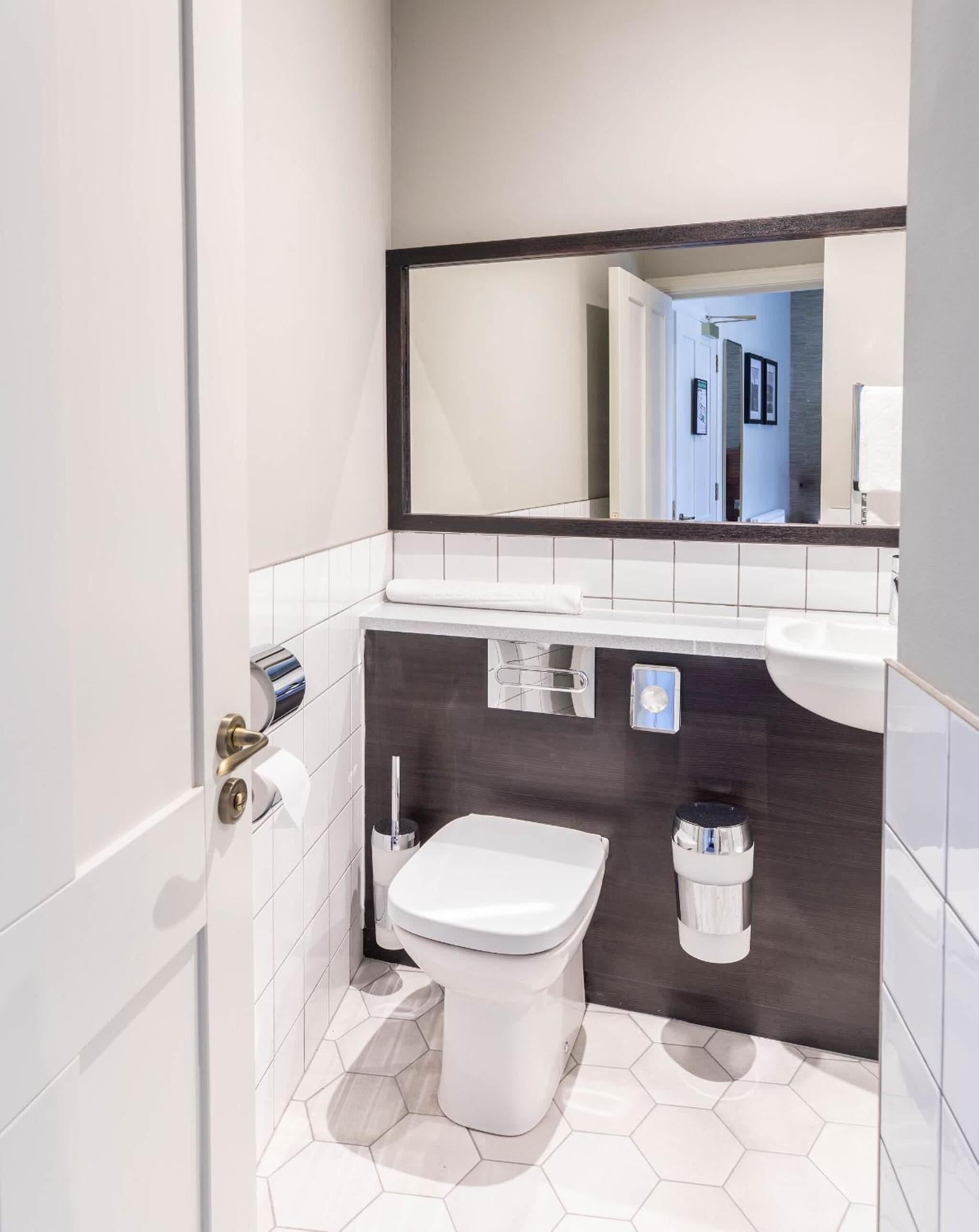 A modern bathroom with a white toilet, a dark tiled wall, a large mirror, and a white sink.