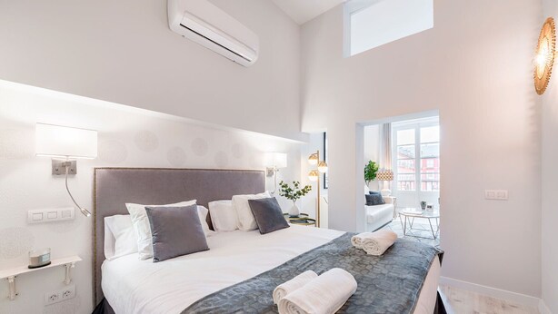 Ein modernes Schlafzimmer mit einem großen Bett, Nachttischen, einer kleinen Sitzecke und Blick auf die Stadt durch ein Fenster.