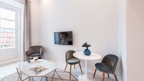 Ein modernes Wohnzimmer mit einem runden weißen Tisch, zwei grauen Stühlen, einer kleinen blauen Vase, einem Flachbildfernseher an der Wand und einer Zeitschrift auf dem Tisch.
