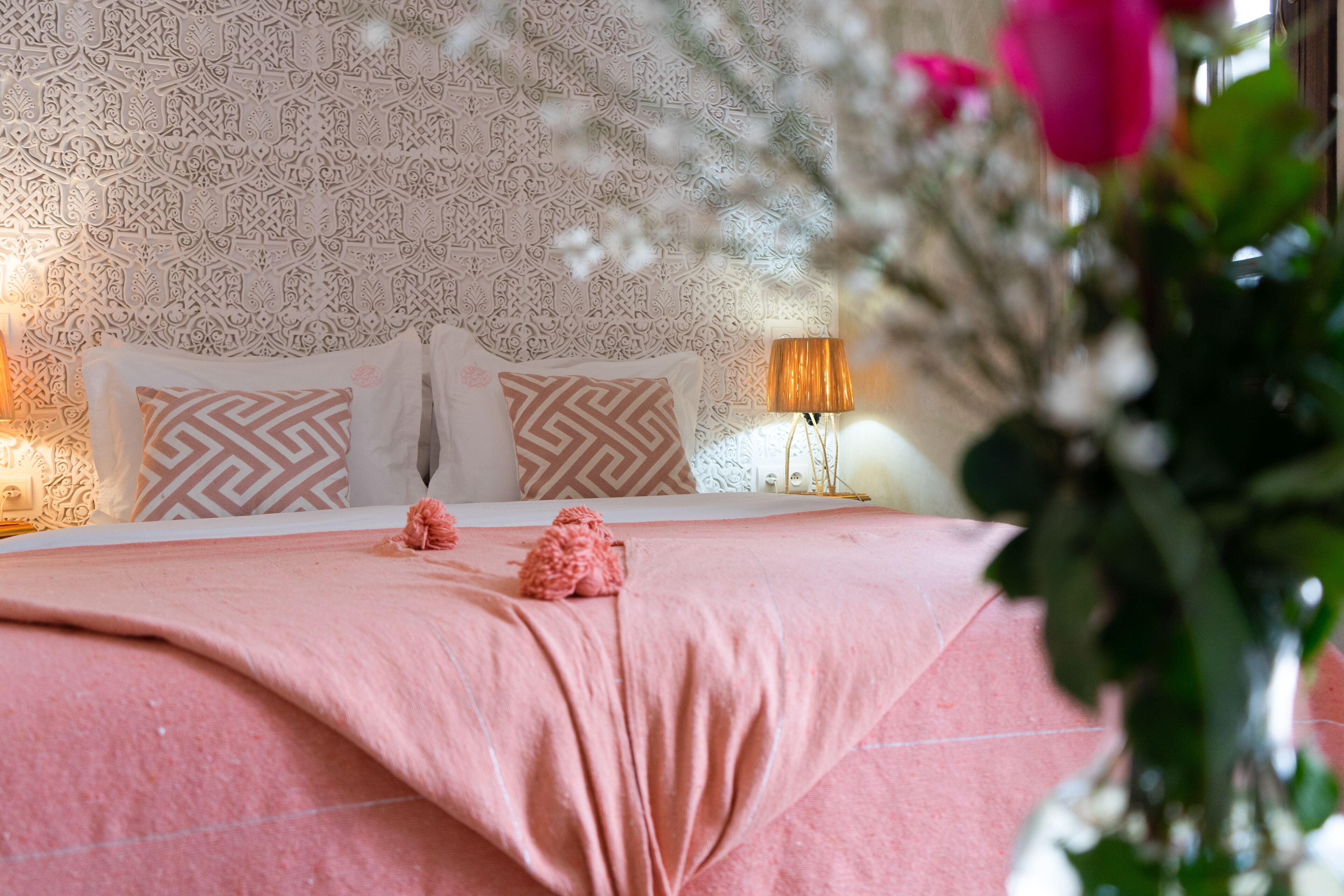 A neatly made bed with patterned pillows, a pink bedspread, and a bedside lamp.