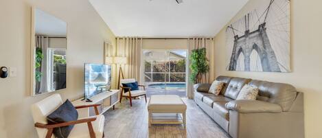 A spacious living room with a ceiling fan, a large window with curtains, a sofa, a chair, and a TV.