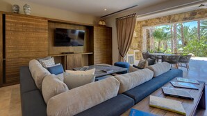 Uma sala de estar moderna com um sofá grande, uma mesa de centro e uma TV de tela plana fixada em uma parede de madeira. É possível ver, por uma grande janela com vista para um jardim tropical, uma área de refeições com mesa e cadeiras.
