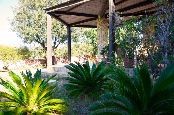 A covered outdoor area with stone pillars, surrounded by lush greenery and trees.