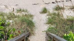 Un sentier bordant la plage et menant à l’océan, agrémenté de rambardes en bois et d’une végétation clairsemée.