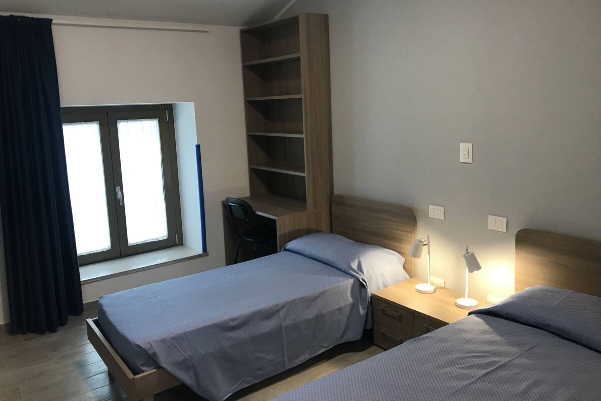 A bedroom with two beds, a desk, a chair, a window with curtains, and a built-in shelving unit.