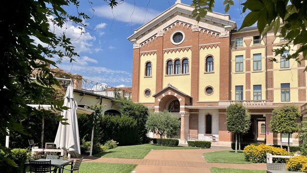 A courtyard with a building featuring a central archway, surrounded by greenery and outdoor seating. - Casa per ferie Don Bosco (Verona)