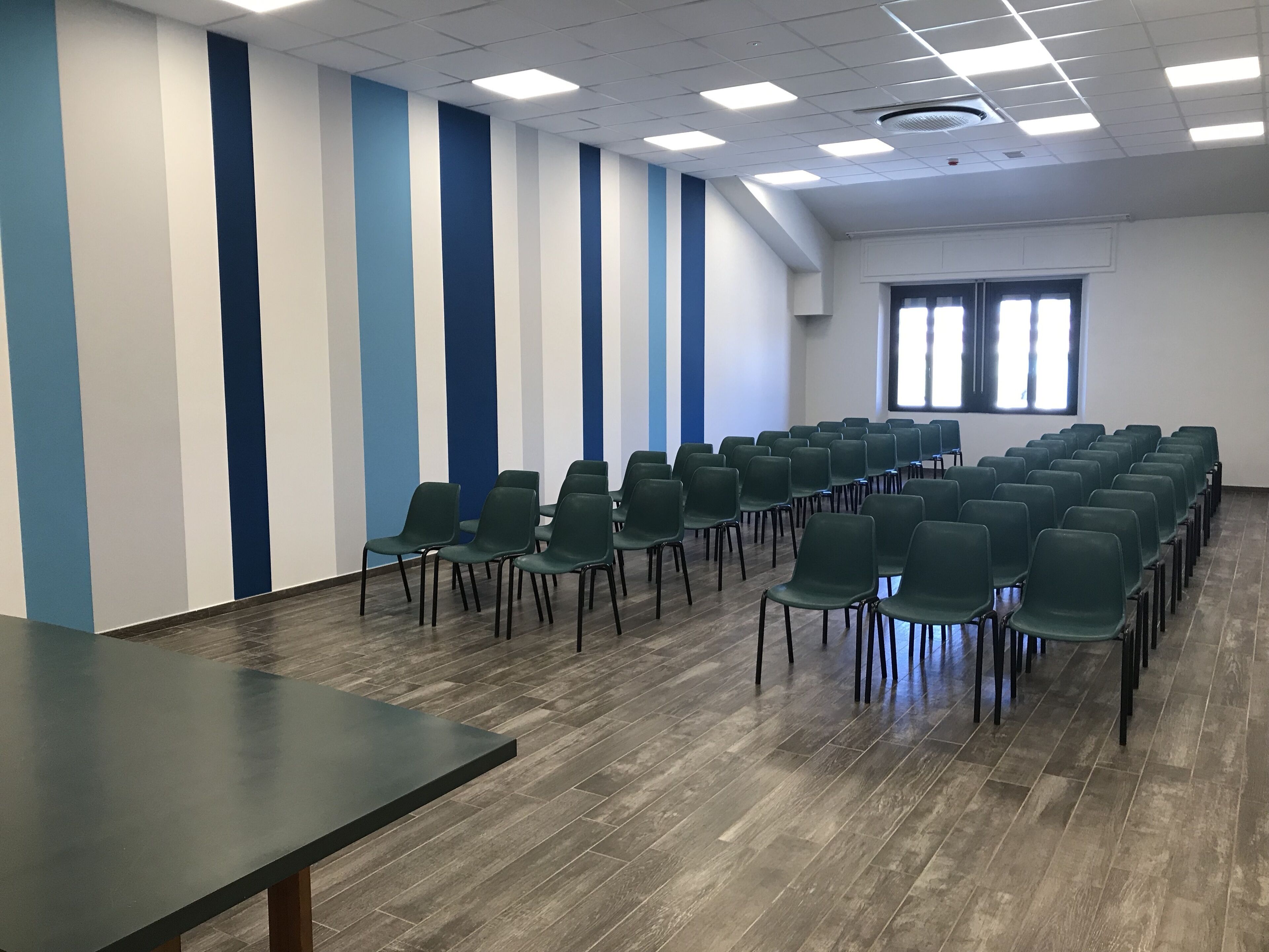 Una sala conferenze con file di sedie verdi, un lungo tavolo e una finestra con persiane.