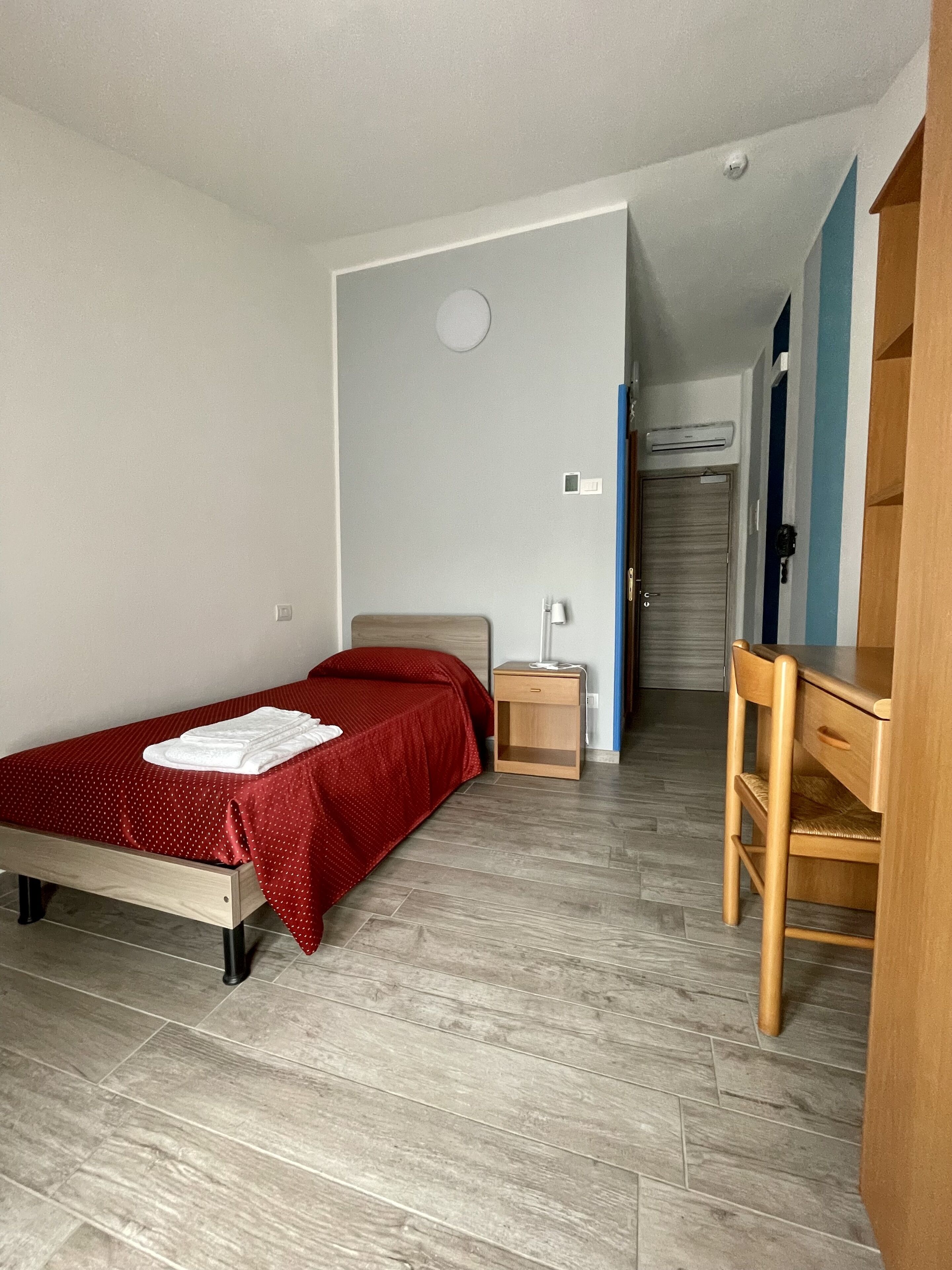 A small, well-lit room with a single bed, a wooden desk, and a chair. There is a small bedside table with a lamp and a power outlet. The room has a tiled floor and a door leading to another room.