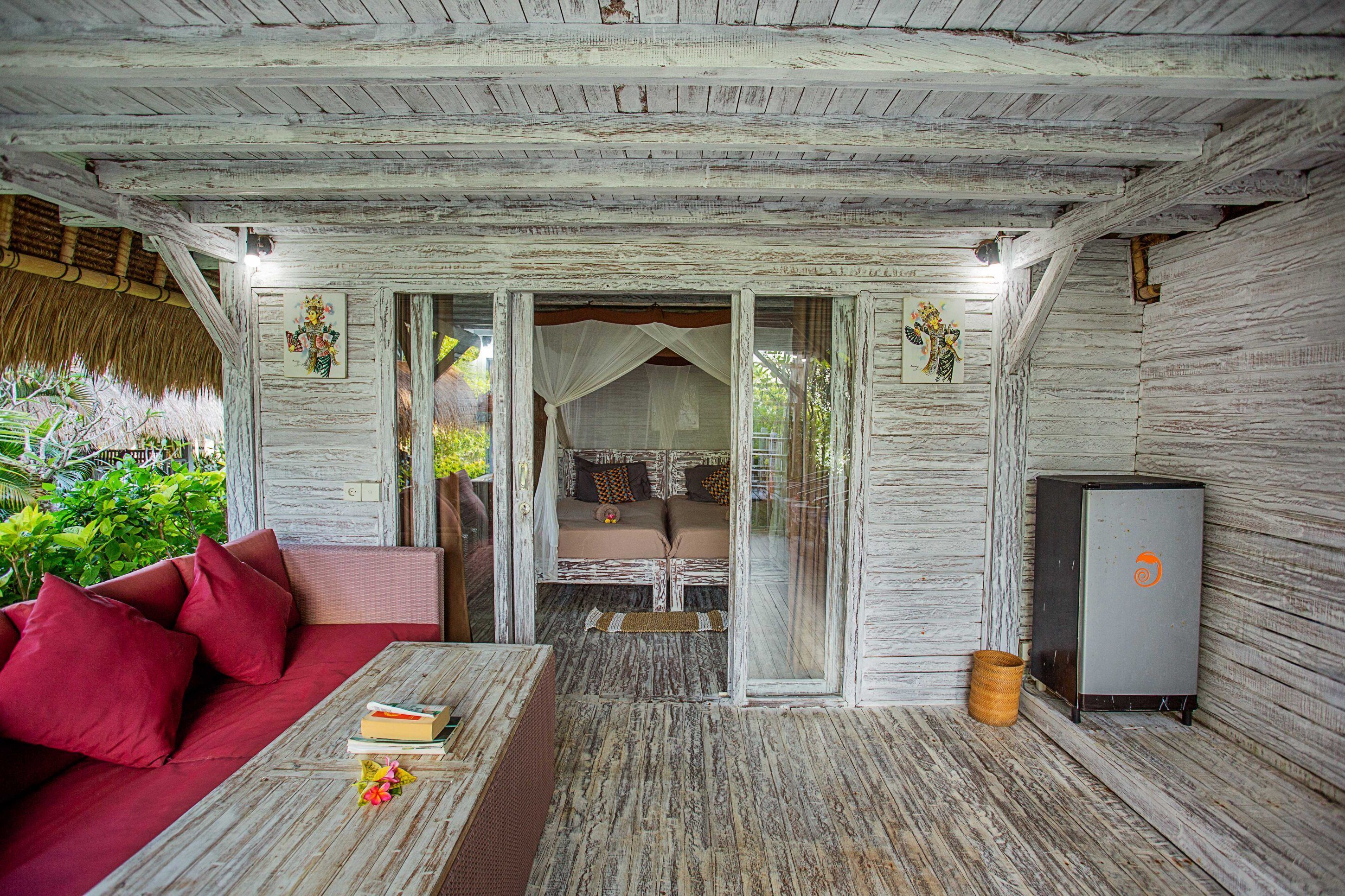 Ein rustikaler Raum mit Holzbalkendecke, einem roten Sofa, einem kleinen Tisch mit Büchern, einem Bett mit Baldachin und einem Kühlschrank.