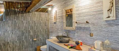 A bathroom with a stone mosaic wall, a wooden ceiling, a large mirror, and a sink with toiletries.