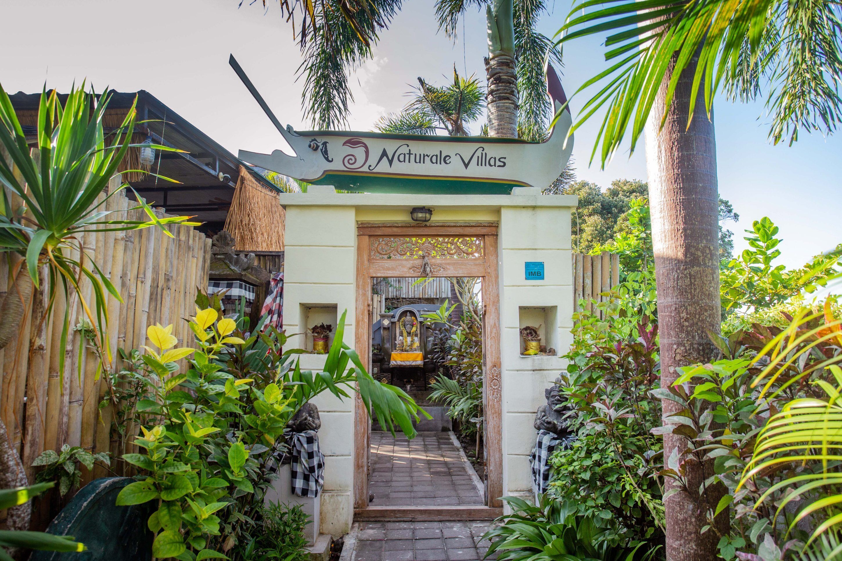 Une entrée aux allures tropicales menant à la propriété baptisée « Naturale Villas », avec un portail en bois, une végétation luxuriante et un ciel dégagé.