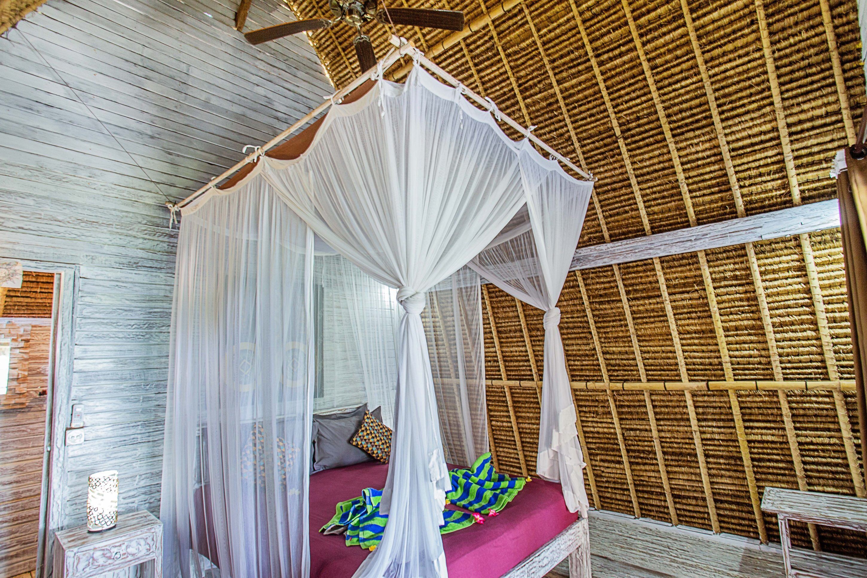 A room with a bed under a white canopy, a wooden ceiling, and a bamboo wall.