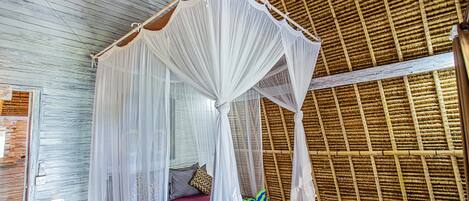 A room with a bed under a white canopy, a wooden ceiling, and a bamboo wall.