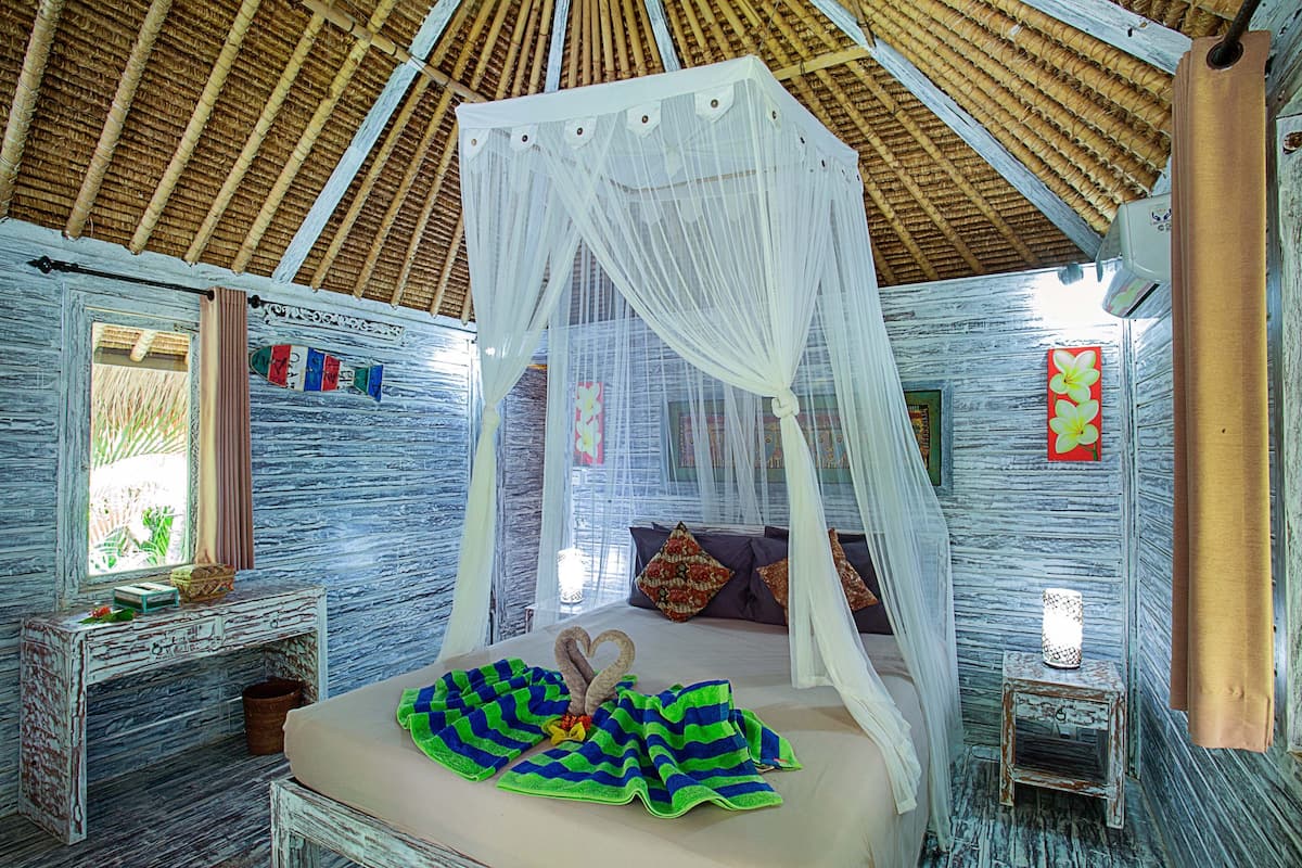 Een kamer met een bed onder een baldakijn, een klein tafeltje met een lamp en een raam met gordijnen.