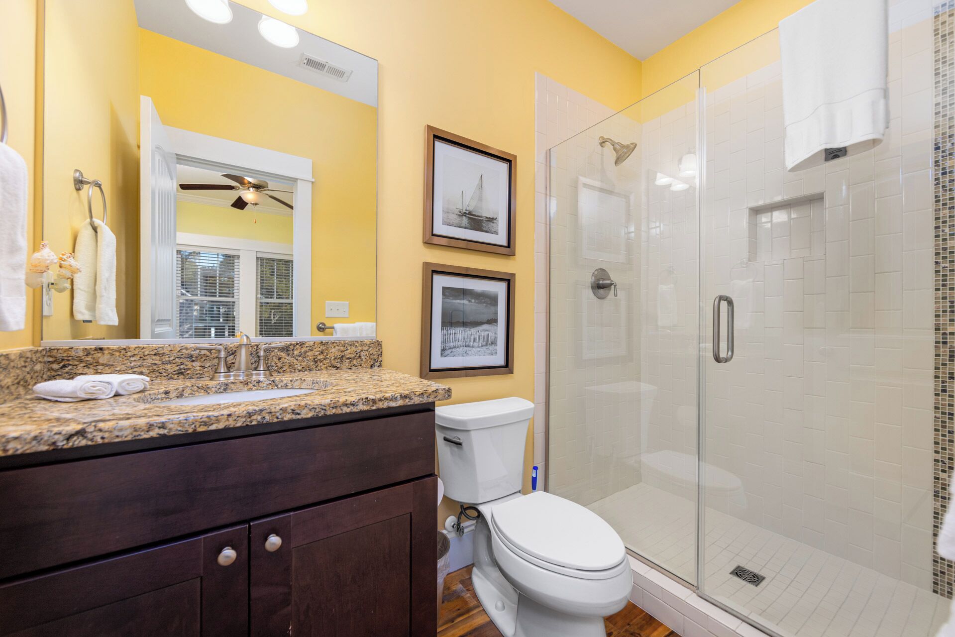 Un baño con encimera de granito, un inodoro, cabina de ducha de vidrio y dos cuadros en la pared.