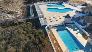 An aerial view of a coastal property with two swimming pools, a beach access bridge, and a residential area with houses.