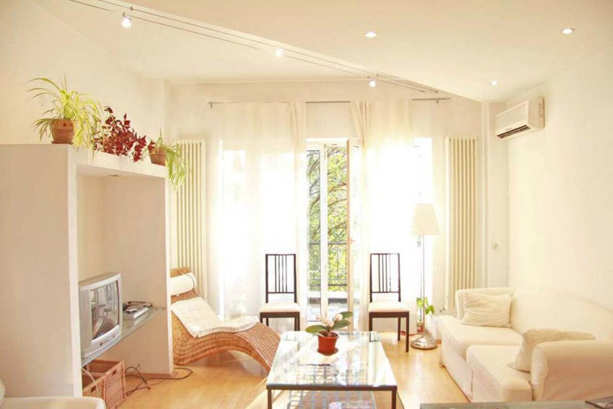 A bright living room with a sofa, chairs, a coffee table, and a TV. There are plants on a shelf and a window with curtains.