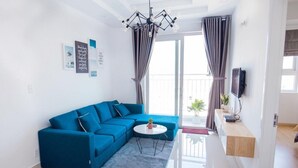 A modern living room with a blue sectional sofa, a small round table, and a chandelier. There are curtains on the windows, a flat-screen TV mounted on the wall, and a potted plant on the windowsill.