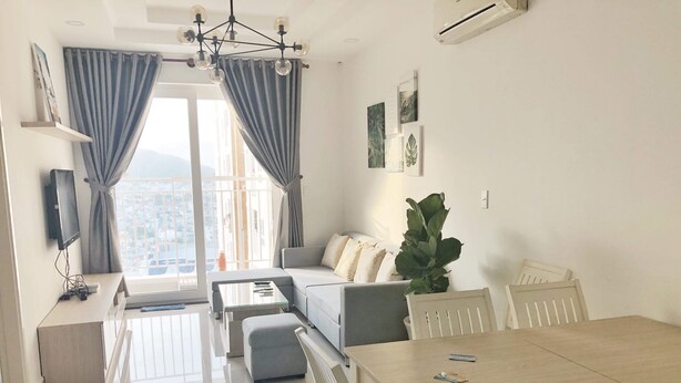 A modern living room with a sofa, a coffee table, a dining table with chairs, a large plant, and a TV mounted on the wall.