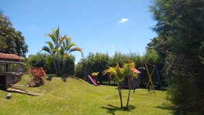 A grassy outdoor area with a playground, including swings and slides, surrounded by trees and a building with a red roof. - Country house in a gated community. (Sorocaba)