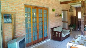 A room with a brick wall, a wooden door with glass panels, a television, a sofa, and a table with a floral tablecloth. - Country house in a gated community. (Sorocaba)