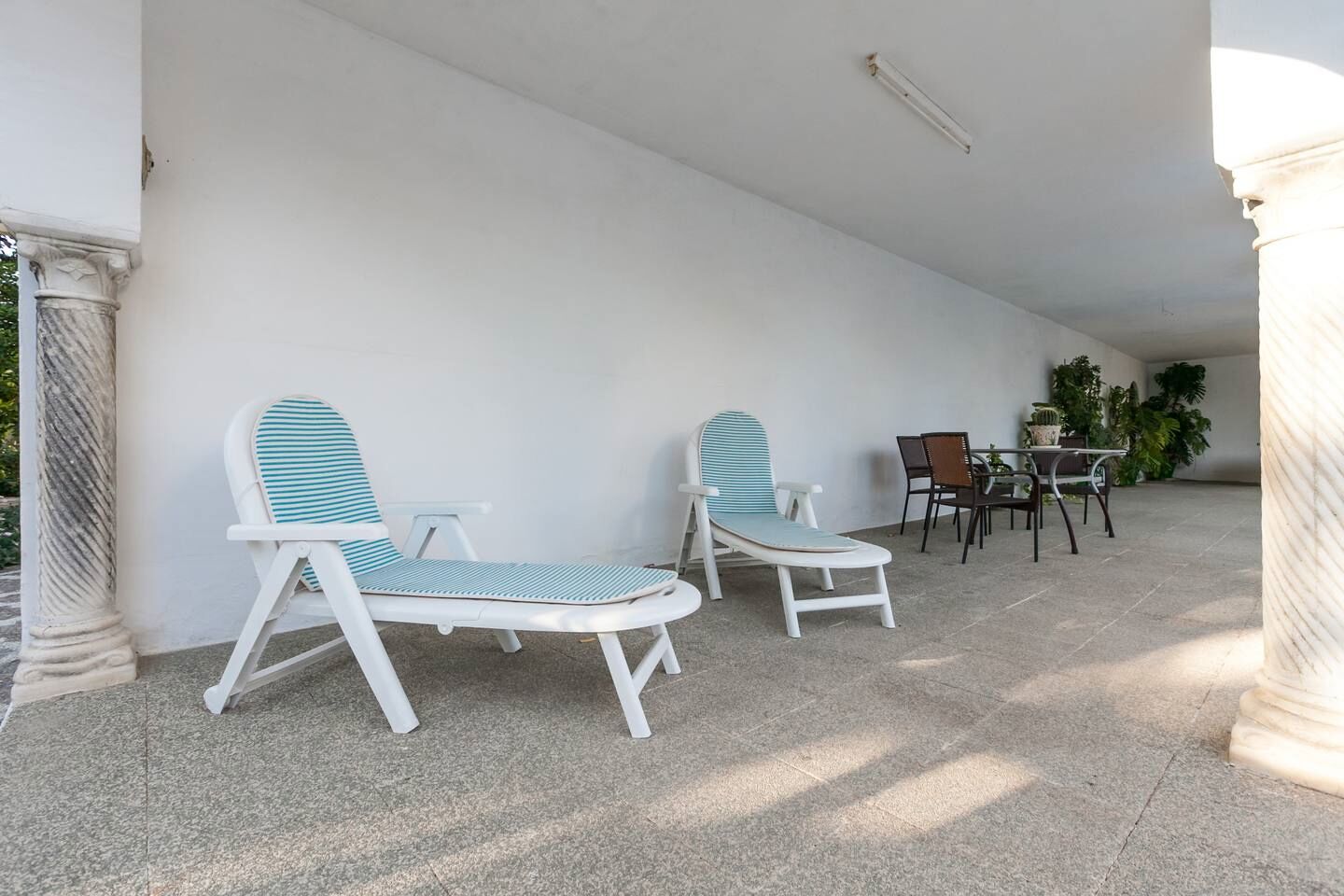 A covered patio area with two white lounge chairs with striped cushions, a small table with chairs, and potted plants.