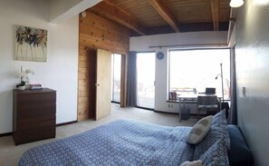 Un dormitorio con una cama grande, techo de madera, un escritorio con una computadora portátil y vistas a través de un ventanal.