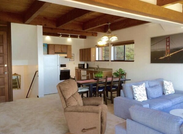 A cozy living room with a beige armchair and a blue sofa, a wooden ceiling, and a kitchen in the background. - 1 bd-1 bth SF Ocean View Rental (Pacifica)