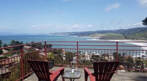 A balcony with two chairs and a table, overlooking a coastal view with a beach and hills.