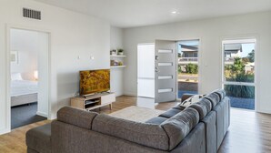 Ett modernt vardagsrum med en grå soffa, en platt-TV, ett litet träbord och en bokhylla. En dörr leder ut till en balkong med utsikt över ett bostadsområde.