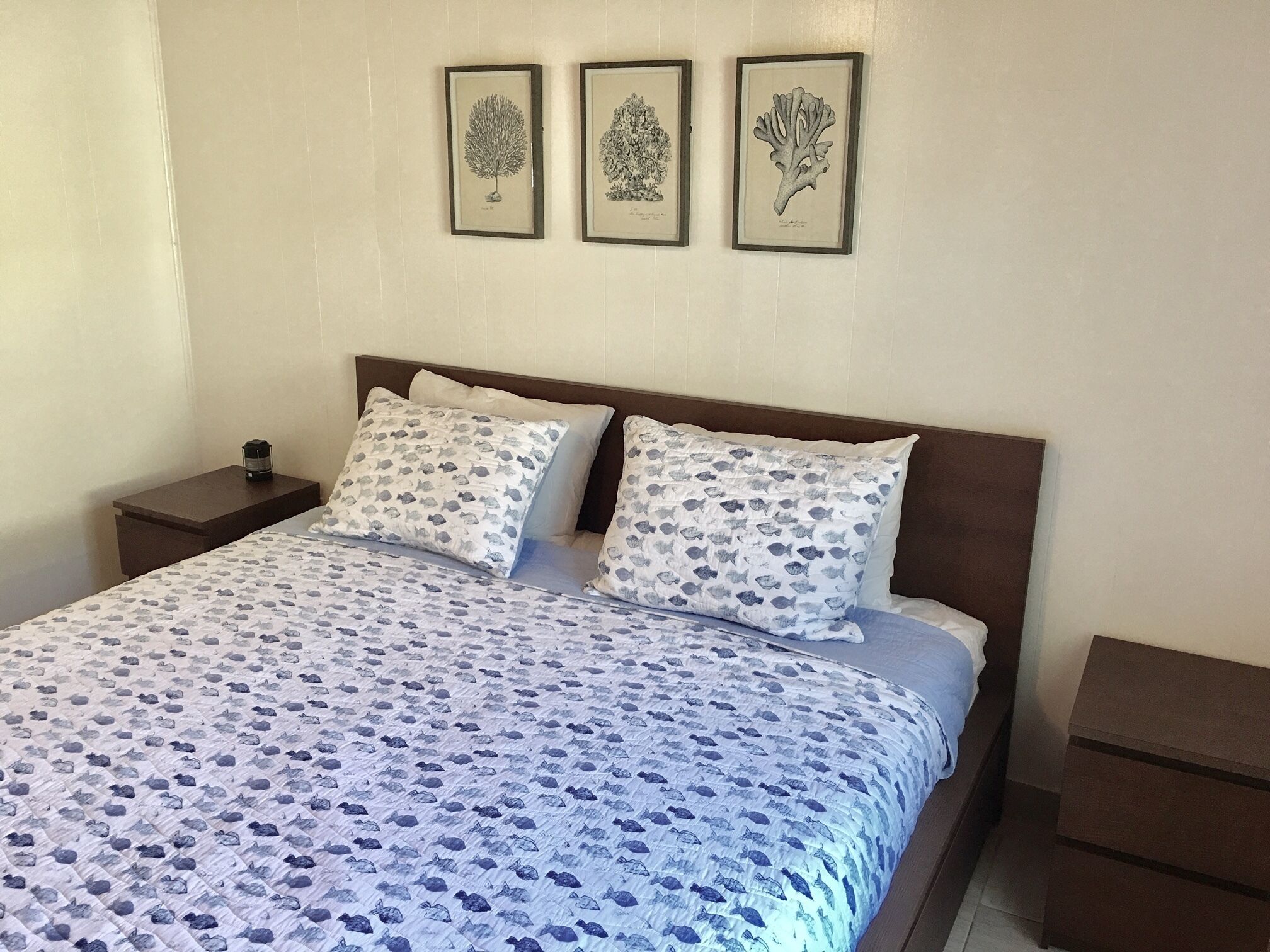 Un lit bien fait, recouvert d’une couvre-lit à motifs, avec deux oreillers décorés de motifs de poissons, et trois illustrations botaniques encadrées accrochées au mur.