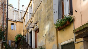 Uma passagem estreita com uma escadaria que leva a um edifício com uma porta de madeira e uma janela com uma persiana. Há plantas em vasos nos degraus e uma bolsa azul no chão.