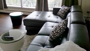 Ein modernes Wohnzimmer mit einem Ecksofa, einem Glastisch und einem großen Fenster mit Vorhängen.