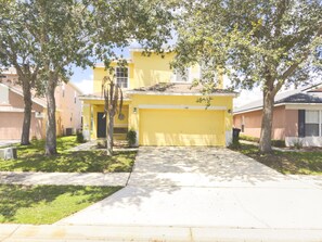 A yellow house with a garage, surrounded by trees and a sidewalk.