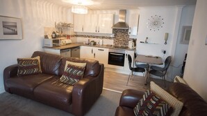 A modern living room with a brown leather sofa, a dining area with a small table and chairs, and a kitchen with white cabinets and a backsplash. - Ideal location for enjoying all of Tenby's attractions (Tenby)
