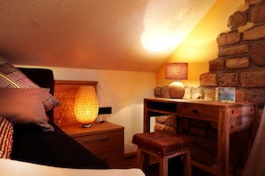 A cozy room with a bed, a wooden desk, a lamp, and a stone wall.