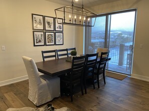Un comedor con una mesa de madera oscura, seis sillas y una araña. Hay cuadros en la pared y un ventanal con vistas al exterior.