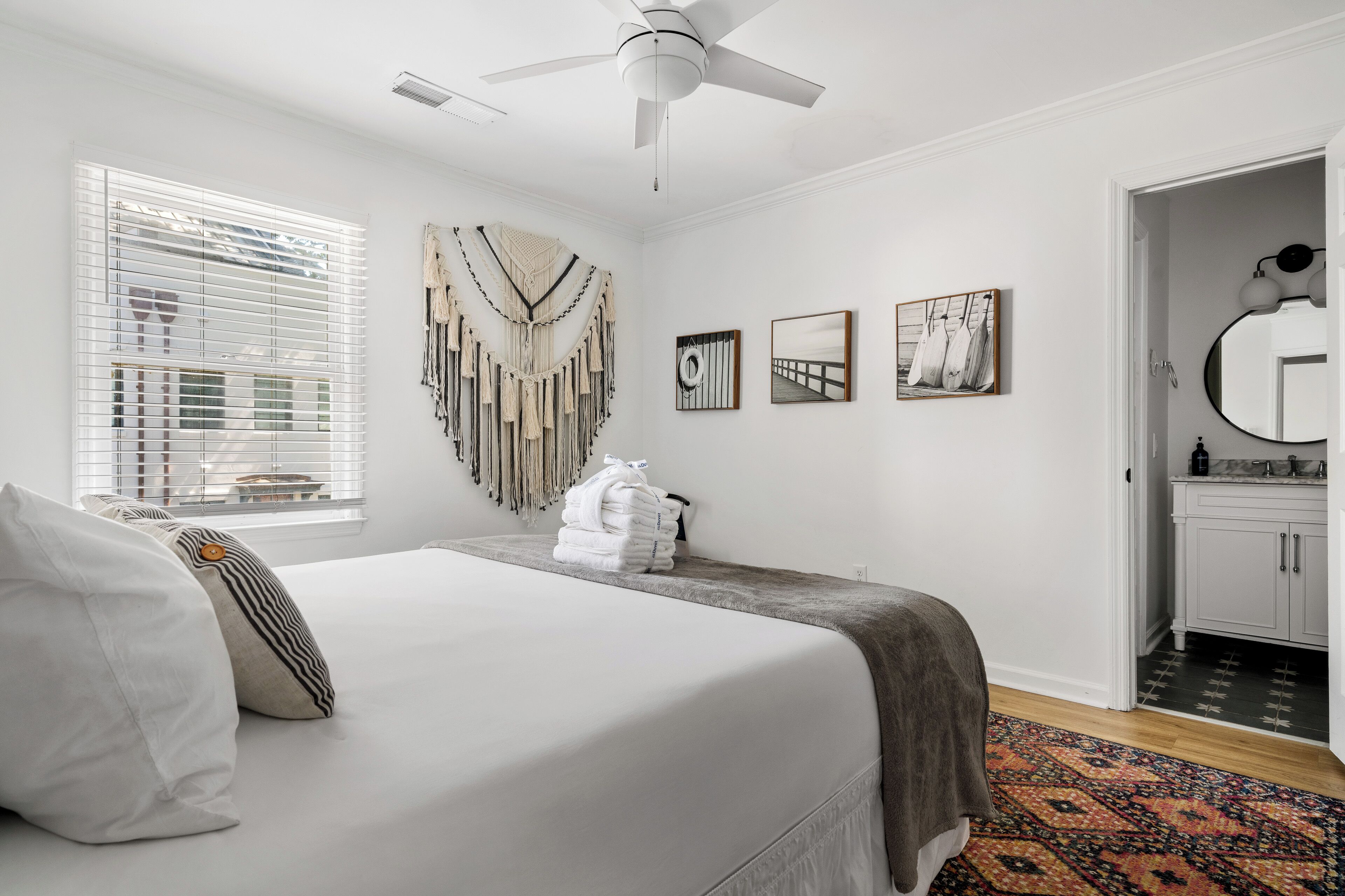 A bedroom with a bed, a ceiling fan, a window with blinds, a wall-mounted tapestry, and a door leading to a bathroom.