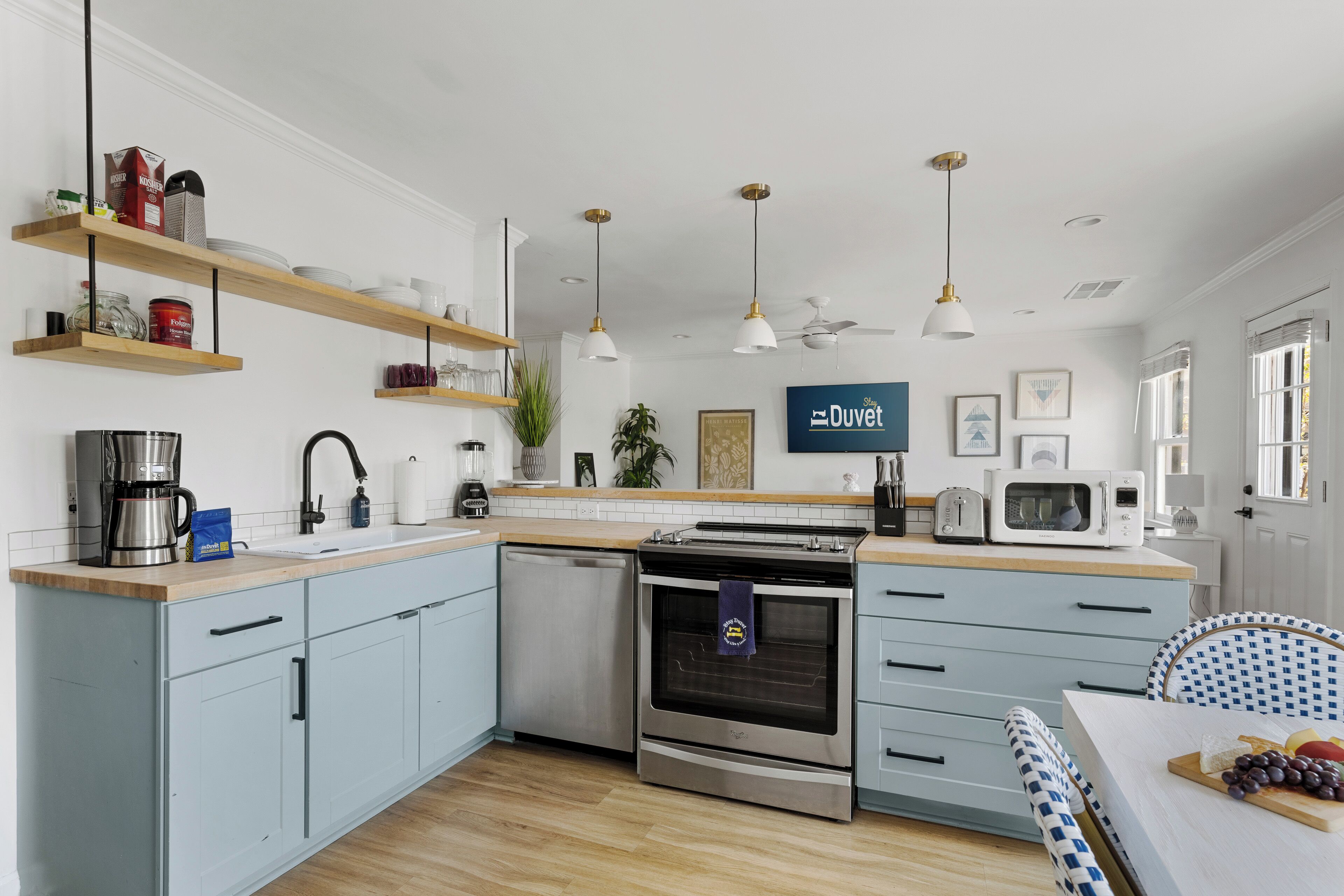 Une cuisine moderne avec des meubles de couleur bleu clair, des appareils en acier inoxydable, des étagères en bois et des luminaires suspendus.