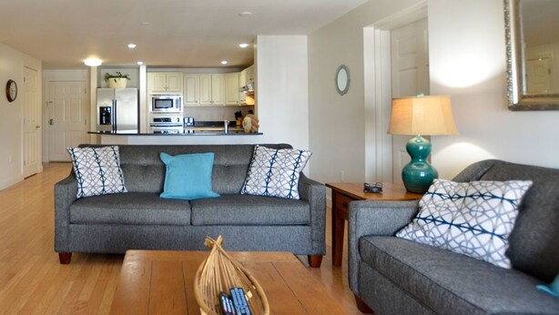 A living room with a grey sofa, a coffee table, and a lamp. There is a kitchen in the background with white cabinets and appliances. - Grand Victorian on the Beach, Summer (Old Orchard Beach)